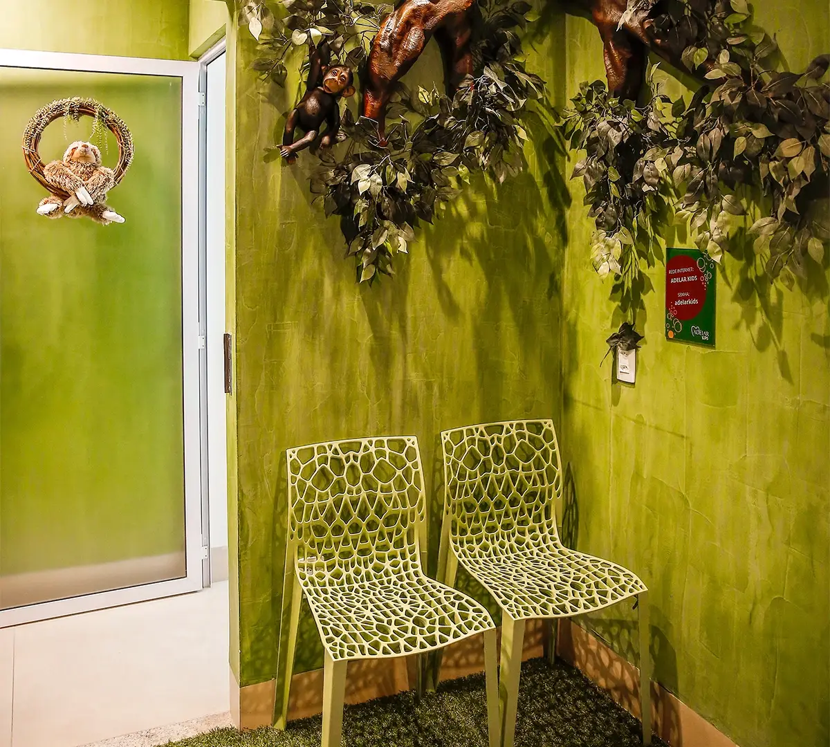 Espaço Adelar Kids: parede verde com galho de árvore, folhagem e bicho de pelúcia, com duas cadeiras infantis e piso de grama sintética